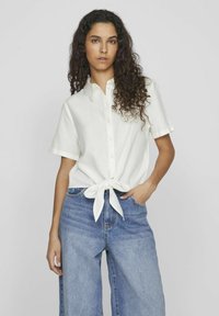 Camicia bianca a maniche corte con chiusura a bottone e dettaglio con nodo davanti, abbinata a jeans a gamba larga blu, su uno sfondo chiaro.