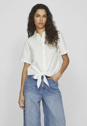 Camicia bianca a maniche corte con chiusura a bottone e dettaglio con nodo davanti, abbinata a jeans a gamba larga blu, su uno sfondo chiaro.