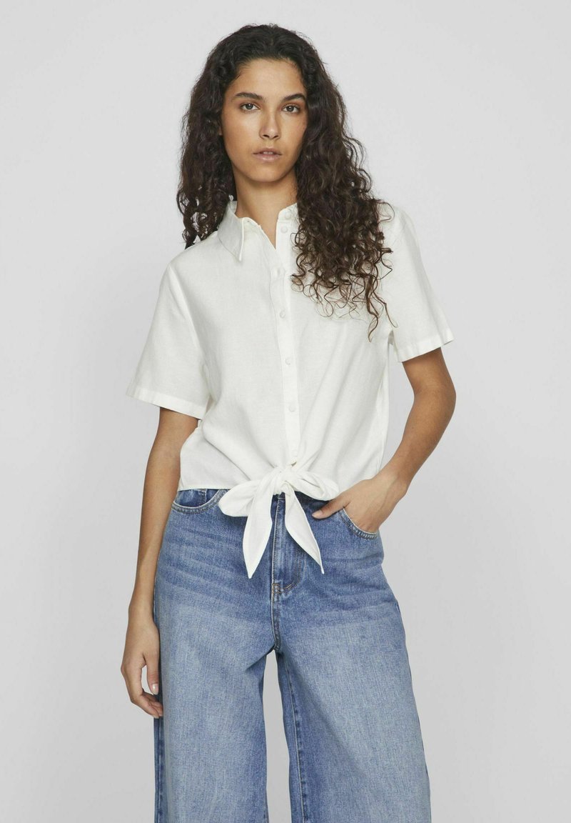 Camicia bianca a maniche corte con chiusura a bottone e dettaglio con nodo davanti, abbinata a jeans a gamba larga blu, su uno sfondo chiaro.