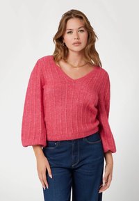 Maglione rosa lavorato a maglia con scollo a V, costine testurizzate e maniche leggermente a sbuffo. Abbinato a jeans blu scuro.