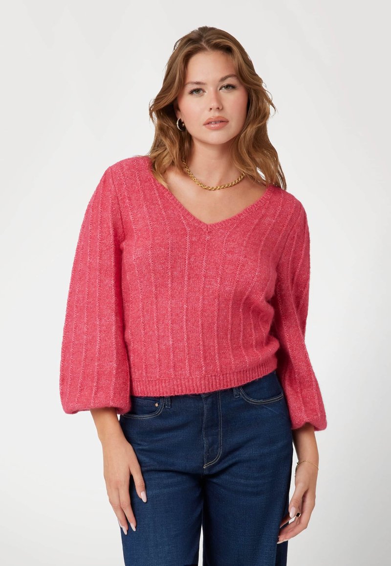Maglione rosa lavorato a maglia con scollo a V, costine testurizzate e maniche leggermente a sbuffo. Abbinato a jeans blu scuro.