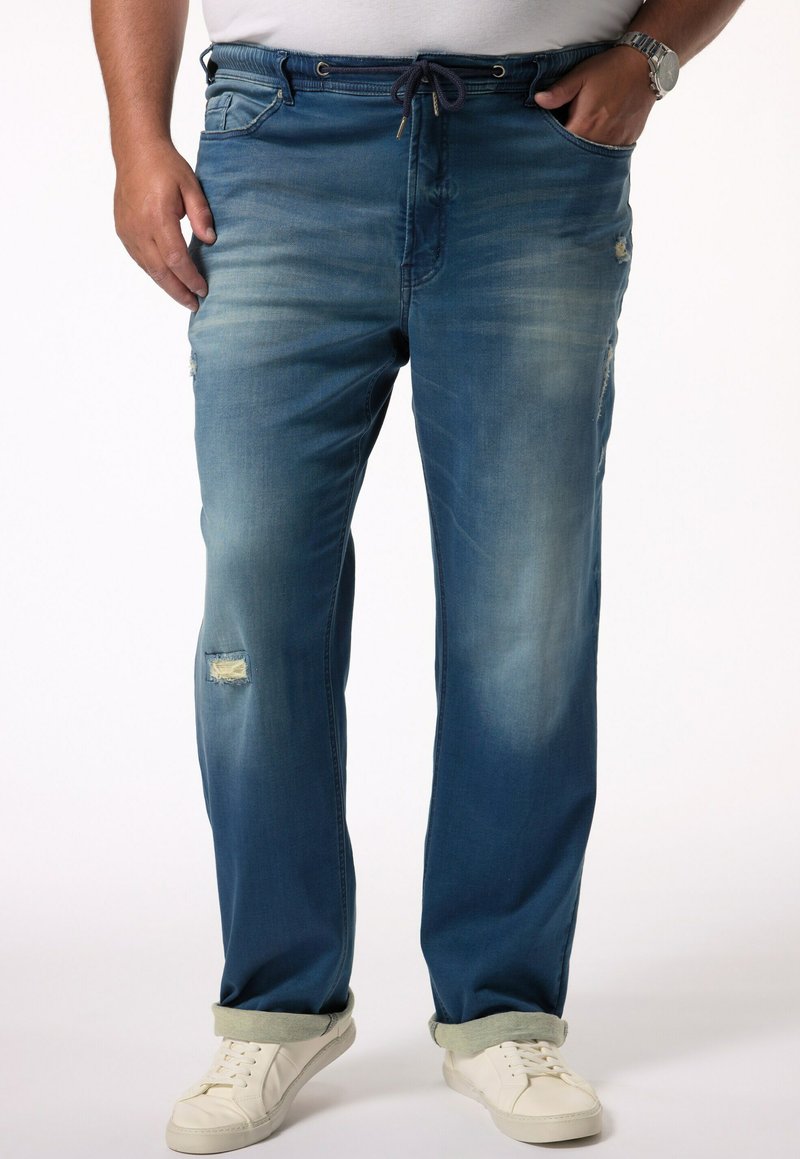 Pantalones jogger de mezclilla en un degradado de azul oscuro a claro, con detalles desgastados, cintura con cordón y puños enrollados, combinados con zapatillas blancas.