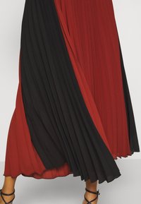 Falda máxima plisada que presenta paneles alternos de tela roja y negra, con una silueta fluida y una textura suave. Se pueden ver tirantes ajustables.