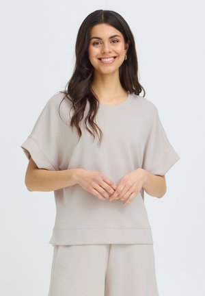 Jeune femme aux longs cheveux bruns souriante, portant un haut ample à manches courtes beige clair et un pantalon assorti, sur un fond uni.
