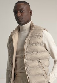 Chaleco aislante beige con patrón de acolchado vertical, cuello alto y bolsillo con cremallera, llevado sobre un suéter de cuello alto beige claro.