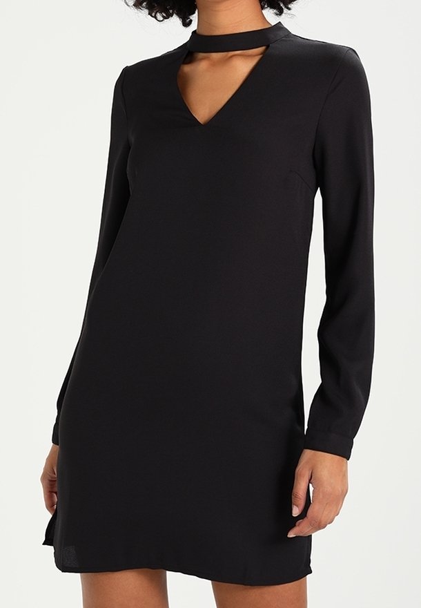 Femme portant une robe noire à manches longues avec un décolleté en V et un détail de collier ras-du-cou, debout devant un fond uni clair.