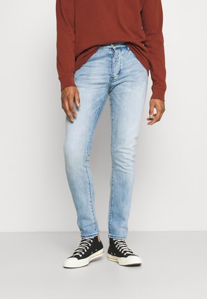 Persoon die lichtblauwe skinny jeans, zwarte hoge sneakers met witte veters en een roestkleurig shirt met lange mouwen draagt, staand.