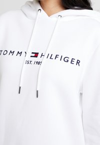 Biała bluza z kapturem, z sznurkiem, na której znajduje się granatowe logo "TOMMY HILFIGER" z czerwonymi i niebieskimi akcentami oraz miękką, gładką teksturą.