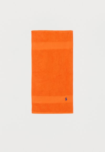 Serviette orange vive avec des bandes texturées et un petit logo brodé bleu dans le coin inférieur droit, sur un fond blanc uni.
