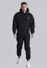 Hoodie di cotone nero con tasca kangaroo e logo dorato, abbinato a pantaloni jogger neri con polsini elastici, indossato con sneakers bianche.