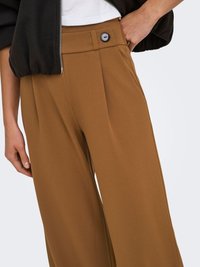 Femme portant un pantalon marron taille haute à jambes larges avec un détail de bouton noir, un haut blanc, et une veste courte noire avec fermeture éclair.