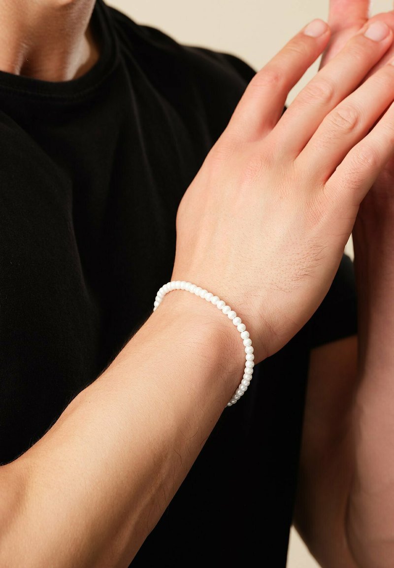 Person trägt ein weißes Perlenarmband am linken Handgelenk, Hände dicht beieinander, bekleidet mit einem schwarzen Kurzarmshirt vor neutralem Hintergrund.