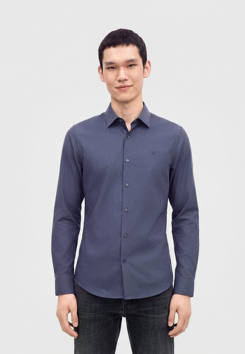 Jeune homme portant une chemise cintrée à manches longues bleu foncé boutonnée et un jean noir, debout devant un fond clair uni.