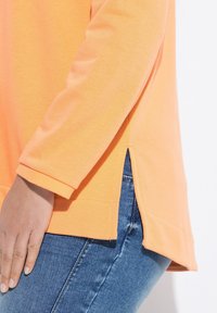 Orange Sweatshirt mit langen Ärmeln, lässiger Passform, seitlichen Schlitzen am Saum und gerippten Bündchen. Kombiniert mit Jeans.