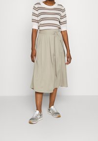 Beige, knielanger Rock mit verstellbarem Bindebund, kombiniert mit einem hellgestreiften kurzärmeligen Oberteil und grauen Sneakers. Glatte Stofftextur.