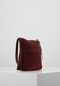 Bordová semišová taška s plochým tvarem, krátkým hnědým koženým popruhem a jednoduchým designem, vybavená jednou vnější kapsou.