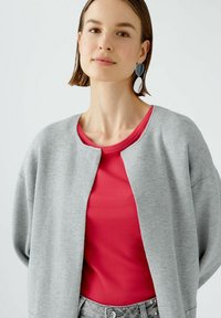 Hellgrauer Cardigan mit rundem Ausschnitt, über einem gerippten rosa Top getragen. Das Model hat kurze Haare und trägt ovale Ohrringe.