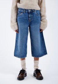 Jasnoniebieskie dżinsowe szerokie spodnie typu culottes z postrzępionym dołem, wysokim stanem i pięcioma kieszeniami, połączone z czarnymi butami i kremowymi skarpetkami w prążki.