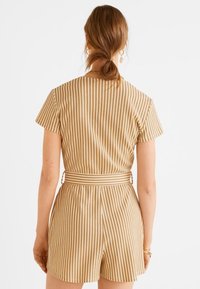 Jumpsuit rayado beige con mangas cortas, un cinturón ceñido en la cintura y tejido texturizado. El diseño presenta rayas horizontales y verticales.