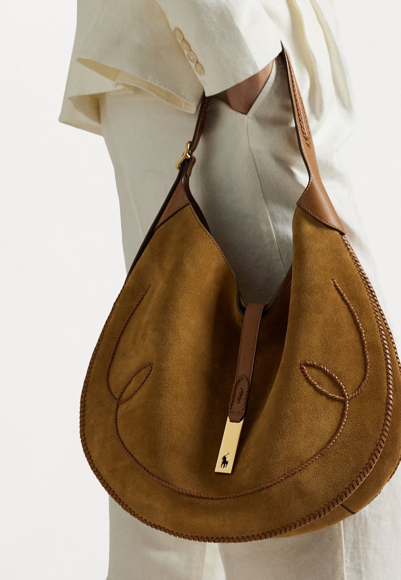 Δερμάτινη τσάντα σε ζεστό καφέ χρώμα με σχήμα χόμπο, διαθέτοντας καμπυλωτά σχέδια ραφής και χρυσό μεταλλικό στοιχείο για κλείσιμο.