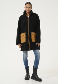 Zwarte fleecejack met hoge kraag, oranje rits en twee beige zakken van leer. Gekoord over blauwe jeans en zwarte enkelboots.