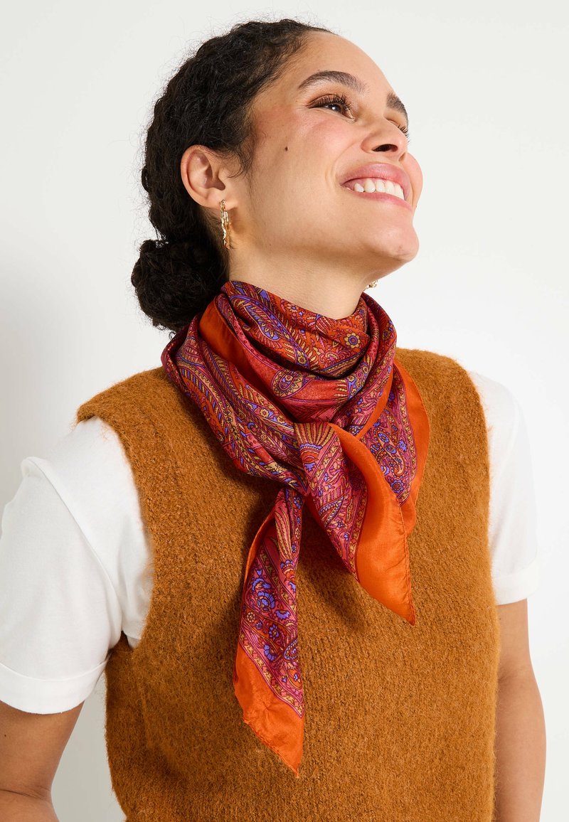 Bufanda de seda en un vibrante color naranja con intrincados patrones paisley en púrpura y rosa, anudada alrededor del cuello, complementada por un chaleco marrón texturizado.