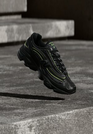 Črn teniški čevelj New Balance z neonsko zelenimi poudarki, ki lebdi nad sivimi betonskimi stopnicami v slabi svetlobi.