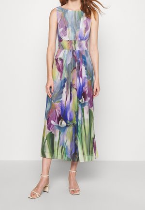 Frau trägt ein ärmelloses, knöchellanges Kleid mit buntem Blumenmuster und Taillendetail, kombiniert mit weißen Sandalen mit Blockabsatz.