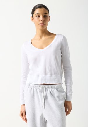 Jeune femme aux cheveux foncés attachés, portant un haut blanc à manches longues avec col en V et un pantalon de jogging gris clair avec cordon de serrage.