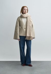 Veste beige en mélange de laine à manches larges, boutonnée croisée, associée à un pull crème, un jean en denim foncé et une écharpe beige à motifs.