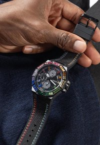 Reloj Guess negro con un bisel octagonal incrustado de gemas multicolores, correa negra texturizada y diales plateados, sostenido en una mano contra un fondo oscuro.