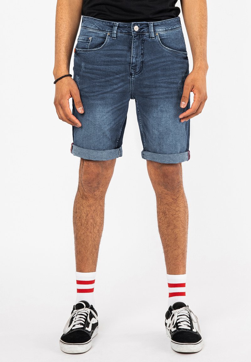 Denim shorts in een medium blauwe wassing, met een klassieke vijf-pocket ontwerp, opgerolde omslagen en een subtiele logo-tag. G gedragen met zwarte sneakers en gestreepte sokken.