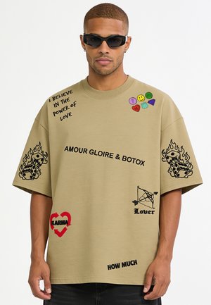 Mann trägt beige oversized T-Shirt mit schwarzen und roten grafischen Drucken, darunter Würfel in Flammen, Textphrasen und bunte kleine Symbole nahe dem Kragen.
