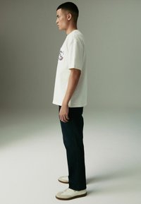 Balta medvilninė marškinėliai su grafiniu spausdinimu kairiajame krūtinės pusėje, derinami su tamsiai mėlynomis džinsais ir baltais sportbačiais su rudos spalvos padais.