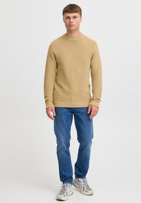 Beige gestrickter Pullover mit geripptem Muster, Rundhalsausschnitt und langen Ärmeln, kombiniert mit blauen Jeans und grauen Sportschuhen.