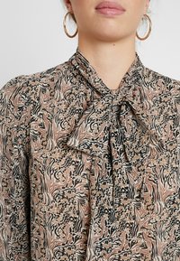 Blusa a maniche lunghe con dettaglio al collo a fiocco, realizzata in tessuto stampato nei colori beige, nero e ruggine, con una finitura morbida e testurizzata.