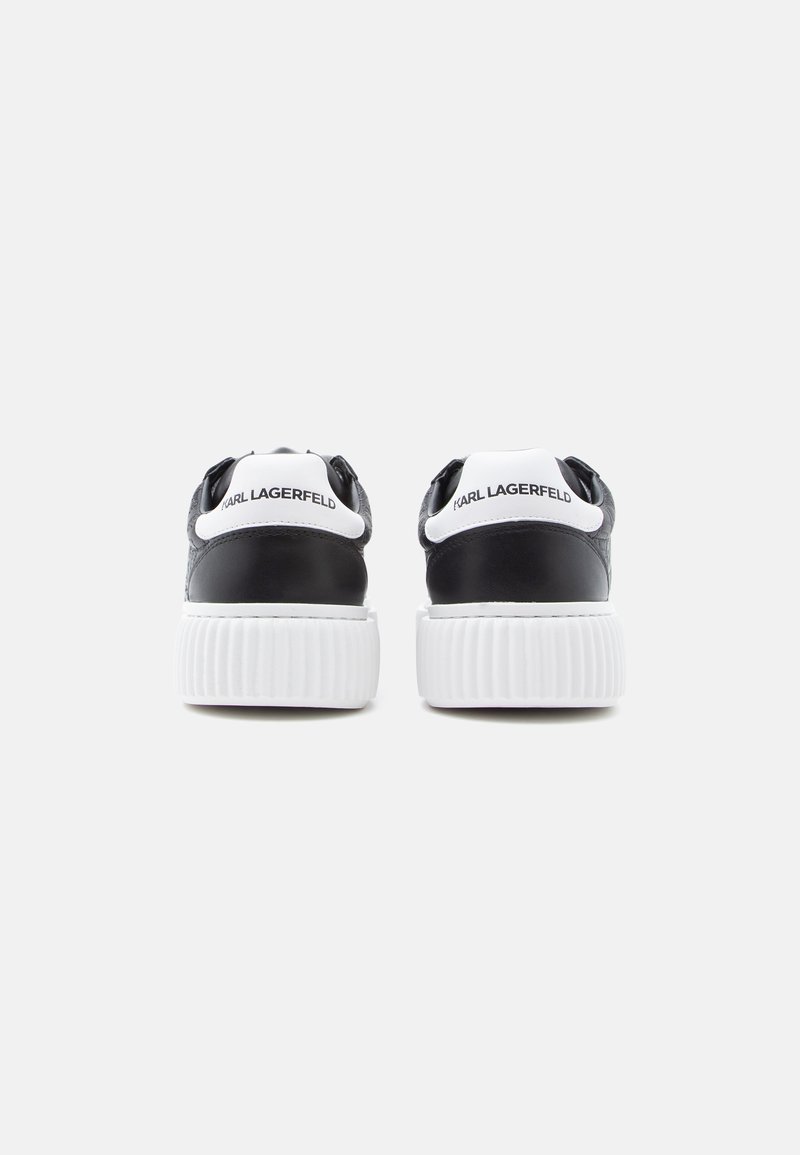 Zapatillas negras de cuero con suela de goma blanca. Presentan un talón texturizado y detalles de marca en blanco en la parte trasera. Forma redondeada.