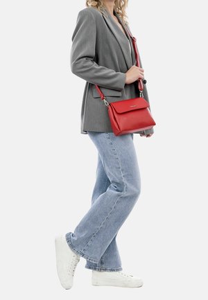 Czerwona skórzana torba na ramię z klapką, srebrnymi elementami i regulowanym paskiem; modelka w szarym żakiecie i jasnoniebieskich dżinsach.
