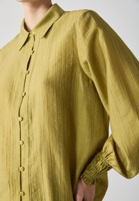 Camicia abbottonata verde oliva con superficie testurizzata, colletto e maniche a palloncino arricciate. Presenta piccoli bottoni sulla parte frontale.
