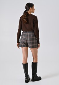 Maglia marrone a maniche lunghe, pantaloni corti a quadri con motivi grigi, neri e arancioni, e stivali neri in pelle al ginocchio con suole spesse.