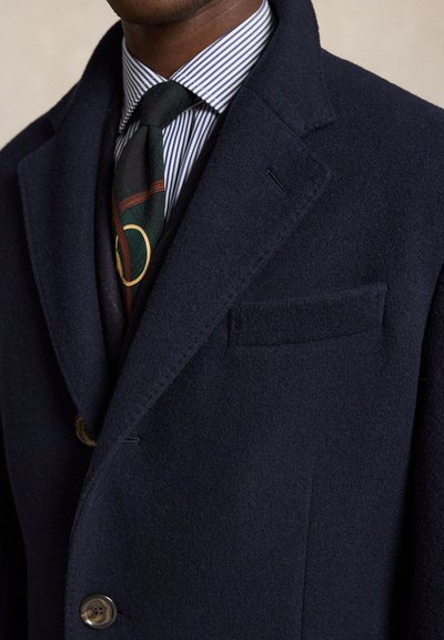 Homme portant un manteau en laine bleu marine par-dessus une chemise à rayures et une cravate sombre à motif doré et rouge.