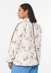 Gewatteerd bloemenjack met een crèmekleurige basis, roze en gele accenten, bruine biezen, lange mouwen en een mandarijnkraag.