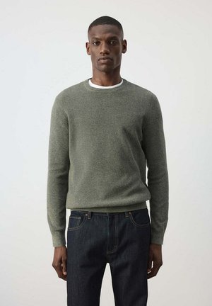 Hombre con pelo corto que lleva un suéter gris ajustado de cuello redondo sobre una camiseta blanca y vaqueros azul oscuro, de pie frente a un fondo claro liso.