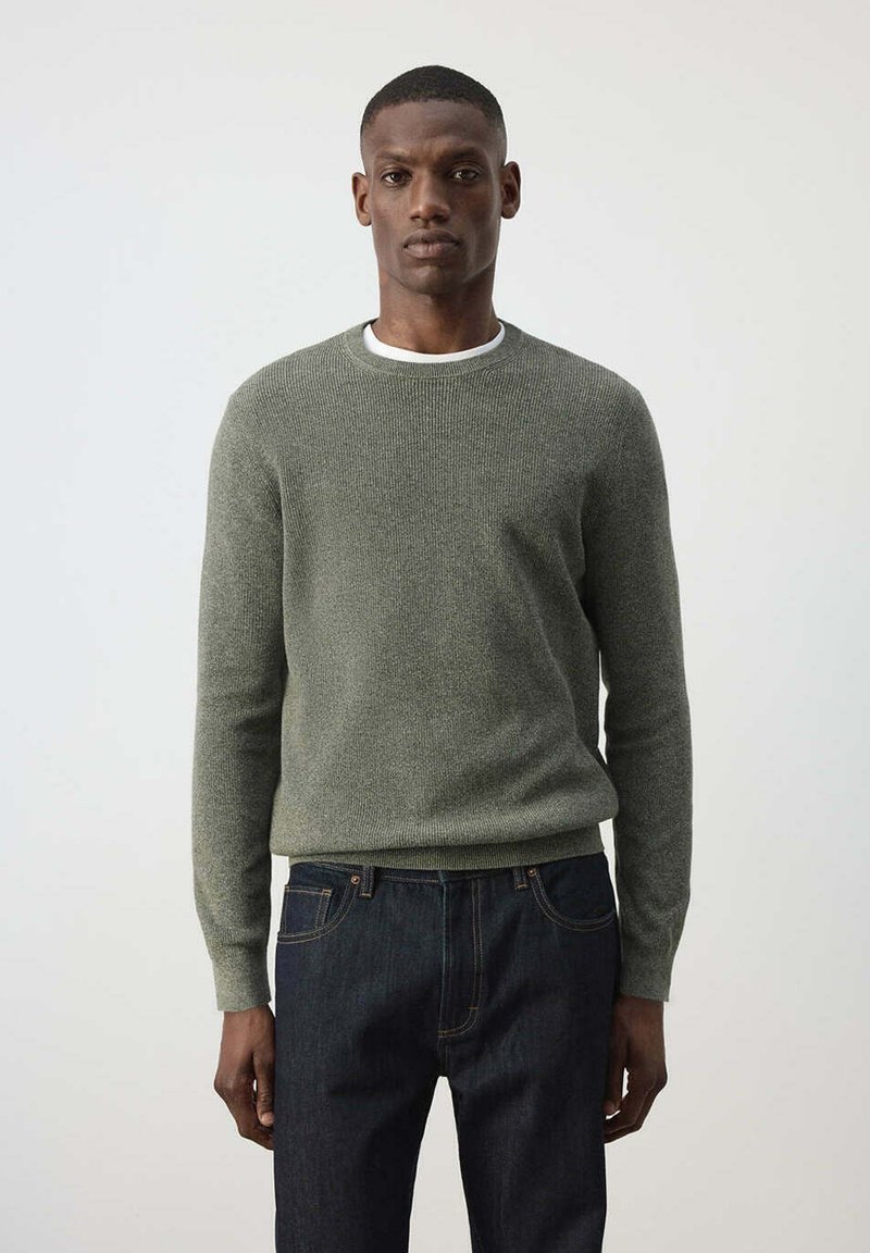 Homme aux cheveux courts portant un pull gris ajusté à col rond par-dessus un maillot blanc et un jean bleu foncé, debout devant un fond clair uni.