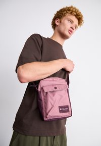 Borsa a spalla in nylon rosa Columbia con patta pieghevole, chiusura a scatto e tracolla regolabile, caratterizzata da un'etichetta sul davanti.