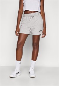 Szürke pamut rövidnadrág, rugalmas derékrésszel és húzózsinórral, fekete adidas logóval a bal oldalon, fehér zoknikkal és cipővel párosítva.