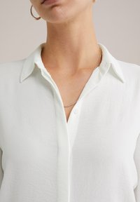 Donna che indossa una camicia bianca con bottoni, una collana di catena argentata e un orecchino a cerchio, mostrata dalla collottola fino alla parte centrale del torso.