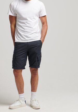 Mann trägt weißes T-Shirt, dunkelblaue Cargoshorts, weiße Kniestrümpfe und weiße Turnschuhe, steht mit Händen in den Taschen.