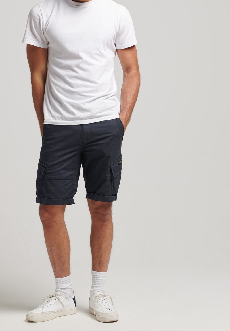 Homme portant un t-shirt blanc, un short cargo bleu foncé, des chaussettes blanches montantes et des baskets blanches, debout les mains dans les poches.