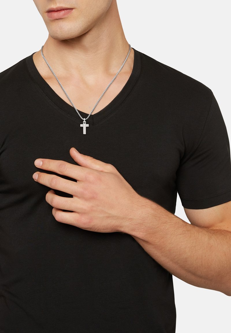 Sort v-hals t-shirt parret med en sølv kæde halskæde med en lille korsvedhæng, der holdes mod brystet. Huden er neutral.